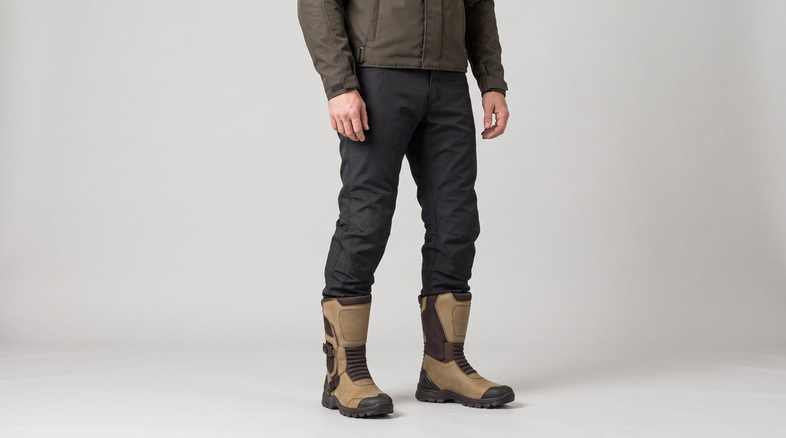 Rider wearing tan and dark brown touring motorcycle boots with dark riding trousers on a light grey studio background