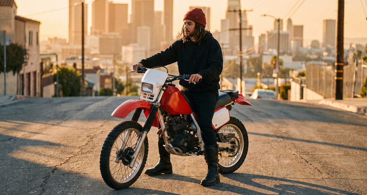 Rider astride an enduro motorcycle on a hill street with the city skyline behind, in tall black touring boots
