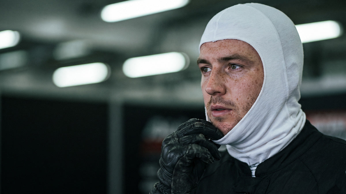 Close portrait of a motorcycle racer pulling on a plain white balaclava in a race garage